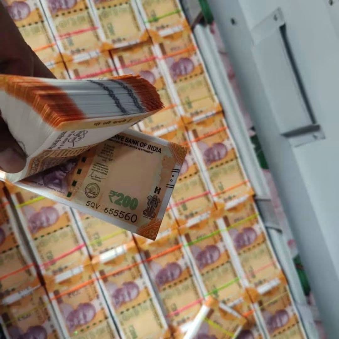 Hand holding a stack of 200 Indian rupee notes over many more cash bundles.