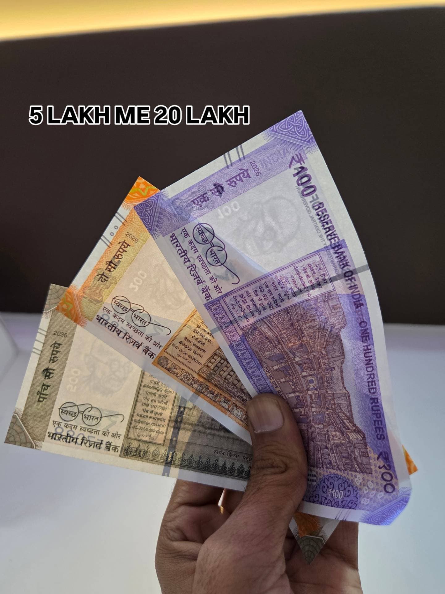 Hand holding Indian Rupee notes 100, 200, 500 with text 5 LAKH ME 20 LAKH.